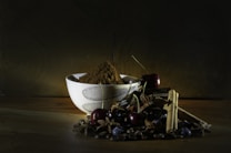 A ceramic bowl filled with powdery brown spices is surrounded by cherries, cinnamon sticks, star anise, coffee beans, and dark berries, all set against a rustic wooden surface and a dark background. The lighting highlights the textures and colors of the ingredients.