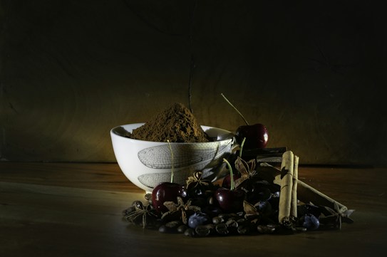 A ceramic bowl filled with powdery brown spices is surrounded by cherries, cinnamon sticks, star anise, coffee beans, and dark berries, all set against a rustic wooden surface and a dark background. The lighting highlights the textures and colors of the ingredients.