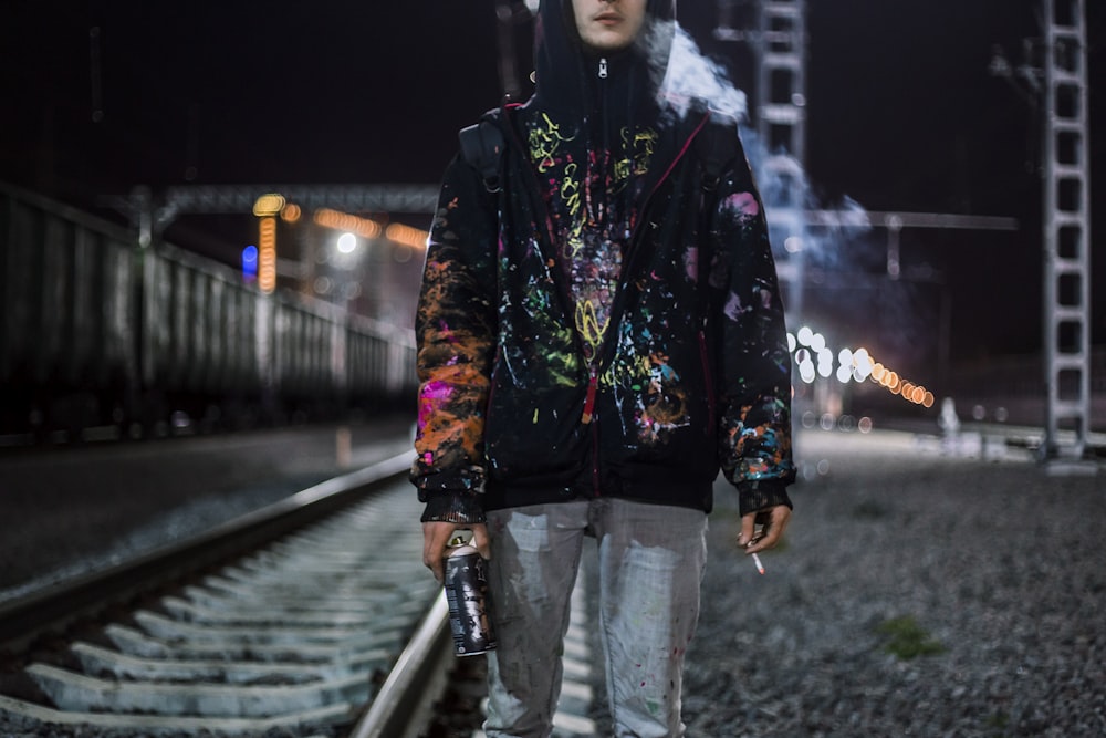 검은 색과 여러 가지 빛깔의 집업 재킷을 입은 남자가 담배를 들고 철도 근처에 서 있습니다.