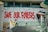 A vibrant graffiti mural with large red letters spells out 'Save Our Forests' on a white wall. The text is adorned with dripping paint, adding to the street art style. To the right, an image of a gorilla holding a spray can is partially visible. A modern walkway with a glass railing is below the mural, where two people are walking. The architecture is sleek and contemporary, featuring clean lines and a blend of glass and concrete.