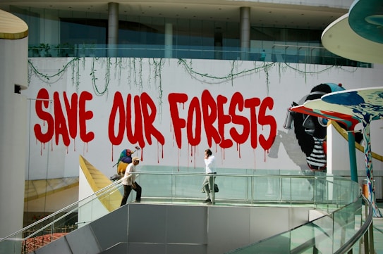 A vibrant graffiti mural with large red letters spells out 'Save Our Forests' on a white wall. The text is adorned with dripping paint, adding to the street art style. To the right, an image of a gorilla holding a spray can is partially visible. A modern walkway with a glass railing is below the mural, where two people are walking. The architecture is sleek and contemporary, featuring clean lines and a blend of glass and concrete.