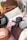 A cozy bed setup features a decorative pillow with the word 'love' embroidered on it, surrounded by various pillows and plush blankets. A dark plush octopus toy rests against the cushions. The bed is under a fabric canopy adorned with hanging star decorations. An old, distressed nightstand sits beside the bed holding a small, warmly lit lamp and some books.