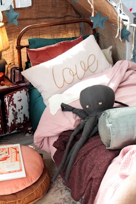 A cozy bed setup features a decorative pillow with the word 'love' embroidered on it, surrounded by various pillows and plush blankets. A dark plush octopus toy rests against the cushions. The bed is under a fabric canopy adorned with hanging star decorations. An old, distressed nightstand sits beside the bed holding a small, warmly lit lamp and some books.