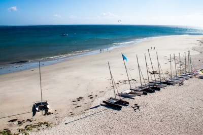 A beach scene with several catamaran sailboats lined up in a row on the sandy shore. The beach is mostly empty, with a few groups of people visible in the distance near the water's edge. The ocean is a deep blue color and stretches out to the horizon. A boat and a kite surfer can be seen in the water further out.