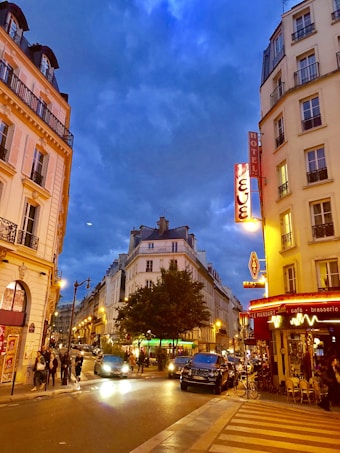 A lively city street scene at dusk with classic European architecture, including several multi-story buildings with balconies. The street is bustling with people walking on the sidewalks and cars driving. There are colorful neon signs for a hotel and a caf&eacute;, which add vibrancy to the scene. The sky is a deep blue with scattered clouds, suggesting an early evening time.