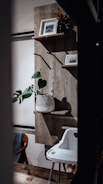A cozy interior space featuring a wooden wall-mounted shelf with two framed pictures and a potted plant. A white, woven basket holds the green plant. Below, a white high chair with a modern design is positioned next to a wooden and fabric chair. The wooden floor adds warmth to the setting.
