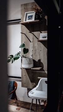 A cozy interior space featuring a wooden wall-mounted shelf with two framed pictures and a potted plant. A white, woven basket holds the green plant. Below, a white high chair with a modern design is positioned next to a wooden and fabric chair. The wooden floor adds warmth to the setting.