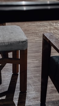 Two wooden chairs with fabric cushions standing on a hardwood floor. The lighting creates distinct shadows, adding contrast to the scene. The floor has a light sheen, indicating a polished surface.