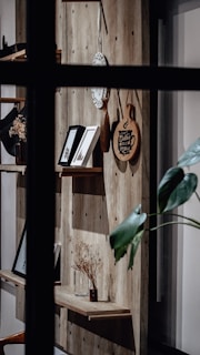A wooden wall-mounted shelving unit is visible, decorated with framed pictures, a decorative plate, cutting boards, a glass vase with dried flowers, and other items. There is also a pothos plant with large green leaves partially in the frame. The scene is viewed through a window with dark, crisscrossing muntins.