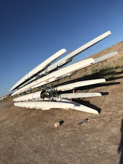 Several rowing boats are stacked on top of each other on a metal rack placed on a dirt surface. The sky is clear with a deep blue hue, and the ground has dry grass patches. The boats are predominantly white with some red accents.