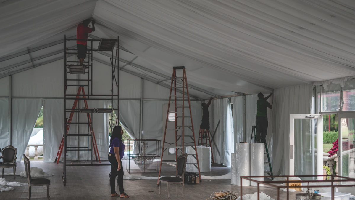 A woman stands in a white event tent watching a crew on ladders hang drapes and light fixtures before an event