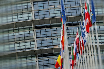 A row of various national flags on tall poles in front of a modern glass building.