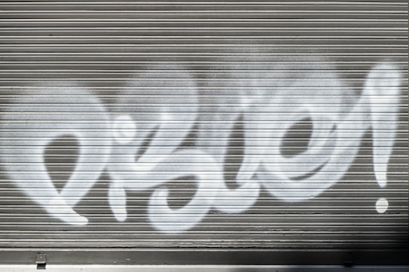 White graffiti text is spray-painted across a metallic gray roller shutter. The letters are bold and stylized, with prominent curves and loops.