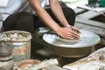 An artist at work, shaping clay on a pottery wheel, with tools and materials arranged nearby.