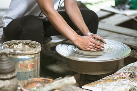 Hands are shaping clay on a spinning pottery wheel, with pottery tools and a paint can nearby. The surrounding area appears to be a rustic, workshop setting, with a focus on the artistic creation of pottery.