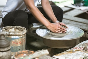 Hands are shaping clay on a spinning pottery wheel, with pottery tools and a paint can nearby. The surrounding area appears to be a rustic, workshop setting, with a focus on the artistic creation of pottery.