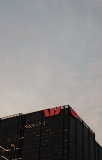A modern glass office building is visible against a cloudy sky backdrop. The structure is characterized by its dark, reflective surfaces and features illuminated interior lights. Red logos are prominently displayed on the top corners of the building.