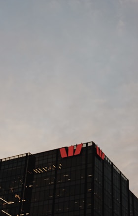 A modern glass office building is visible against a cloudy sky backdrop. The structure is characterized by its dark, reflective surfaces and features illuminated interior lights. Red logos are prominently displayed on the top corners of the building.