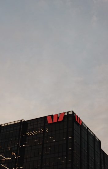 A modern glass office building is visible against a cloudy sky backdrop. The structure is characterized by its dark, reflective surfaces and features illuminated interior lights. Red logos are prominently displayed on the top corners of the building.