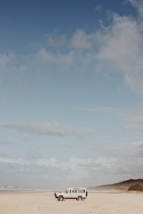 A solitary white off-road vehicle is parked on a wide, sandy beach beneath a vast sky. The horizon is lined with gentle waves, and the sky is partially cloudy with soft, diffused lighting. Distant greenery is visible at the far right edge of the beach.
