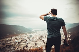 A person stands on a hill overlooking a cityscape, with mountains and a body of water in the background. The individual is holding a camera in one hand and touching their head with the other, seemingly taking in the view.