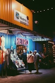 A retro-themed shop with a neon sign reading 'Retro' is adorned with a bright yellow panel above it. The storefront displays various vintage items, including clothing and magazines. People are gathered outside, some engaging in conversation. The setting is warmly lit, creating a cozy ambiance.
