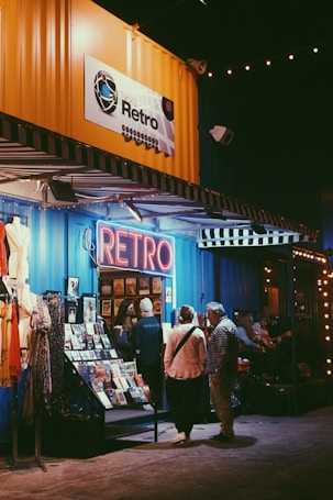 A retro-themed shop with a neon sign reading 'Retro' is adorned with a bright yellow panel above it. The storefront displays various vintage items, including clothing and magazines. People are gathered outside, some engaging in conversation. The setting is warmly lit, creating a cozy ambiance.