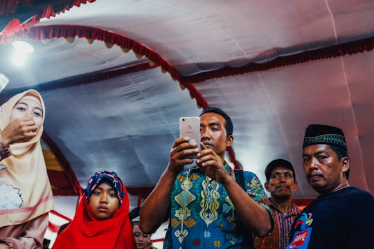 A diverse group of people gathered around a smartphone, engaging with the ummahone app in a warmly lit mosque setting.