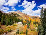 A picturesque mountain landscape featuring a cabin surrounded by dense evergreen trees. The mountains in the background are partly covered in autumn foliage, displaying vibrant orange and yellow colors. The sky above is bright with a mix of puffy white clouds scattered across a deep blue sky.