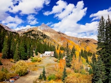 A picturesque mountain landscape featuring a cabin surrounded by dense evergreen trees. The mountains in the background are partly covered in autumn foliage, displaying vibrant orange and yellow colors. The sky above is bright with a mix of puffy white clouds scattered across a deep blue sky.
