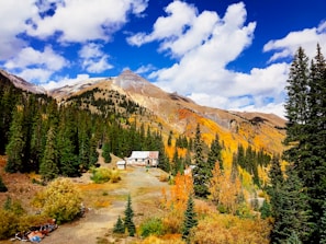 A picturesque mountain landscape featuring a cabin surrounded by dense evergreen trees. The mountains in the background are partly covered in autumn foliage, displaying vibrant orange and yellow colors. The sky above is bright with a mix of puffy white clouds scattered across a deep blue sky.