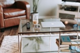 A modern living room scene with a glass coffee table. On the table there is a laptop, a smartphone, a glass jar containing a drink, and two remote controls. In the background, there is a brown leather chair, a white fluffy rug on the wooden floor, a television on a white stand, and a potted plant.