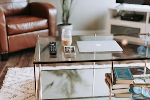 A modern living room scene with a glass coffee table. On the table there is a laptop, a smartphone, a glass jar containing a drink, and two remote controls. In the background, there is a brown leather chair, a white fluffy rug on the wooden floor, a television on a white stand, and a potted plant.