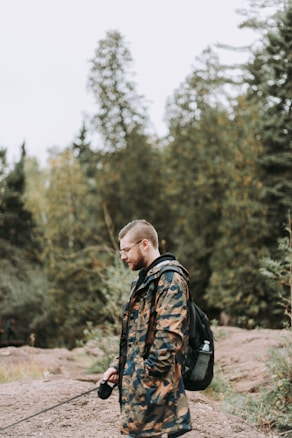 A person wearing a camouflage jacket stands outdoors with a camera in hand, surrounded by dense forest. The individual has light-colored hair and is equipped with a backpack, appearing to be on a nature walk or hike. The background consists of tall trees with green foliage, and the ground is rocky and earthy.