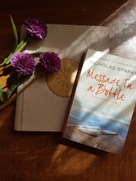 A book titled 'Message in a Bottle' by Nicholas Sparks is positioned on a wooden surface next to a notebook with a decorative emblem. Three purple flowers with green stems are placed on the notebook. Sunlight streams in from an angle, casting shadows and highlighting the textures of the objects.
