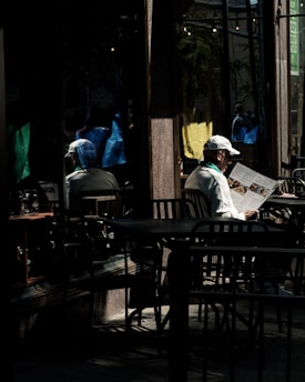 A person sits alone at an outdoor café table, reading a newspaper. The setting is dimly lit with shadows cast by nearby structures. The person wears a white cap and jacket, adding to the contrast against the darker background. Reflections in the café’s glass windows capture vibrant colors from the street, including blue, yellow, and green.