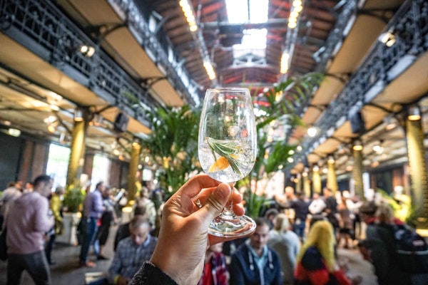 A hand holds a clear glass containing a cocktail with a slice of citrus and a sprig of rosemary, set against the backdrop of a bustling indoor venue with high ceilings and metal beams. The setting appears to be lively, with numerous people mingling and standing around, surrounded by tall green plants.