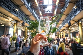 A hand holds a clear glass containing a cocktail with a slice of citrus and a sprig of rosemary, set against the backdrop of a bustling indoor venue with high ceilings and metal beams. The setting appears to be lively, with numerous people mingling and standing around, surrounded by tall green plants.