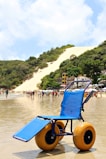 A blue beach wheelchair with large yellow wheels is positioned on a sandy shoreline. People are walking in the background near the water, and a large sand dune covered with patches of greenery is visible behind a row of beach houses.