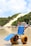 A blue beach wheelchair with large yellow wheels is positioned on a sandy shoreline. People are walking in the background near the water, and a large sand dune covered with patches of greenery is visible behind a row of beach houses.