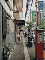 A sidewalk scene in an urban area features a parking payment machine in the foreground, a neatly tiled path with tactile paving, and a variety of plants along the edge. Buildings line the left side, with a canopy extending outwards, and above is a cluster of utility cables. A signage pole indicates parking payment. Several parked motorcycles and cars are visible along the street, with a couple of people walking and sitting in the background. The atmosphere is typical of a bustling city street.