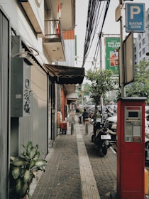 A sidewalk scene in an urban area features a parking payment machine in the foreground, a neatly tiled path with tactile paving, and a variety of plants along the edge. Buildings line the left side, with a canopy extending outwards, and above is a cluster of utility cables. A signage pole indicates parking payment. Several parked motorcycles and cars are visible along the street, with a couple of people walking and sitting in the background. The atmosphere is typical of a bustling city street.