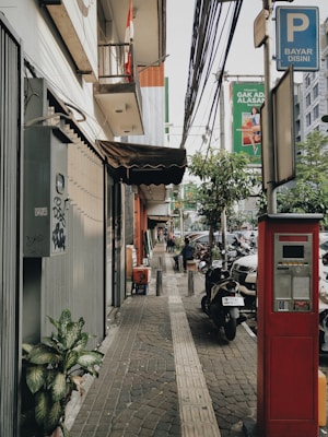 A sidewalk scene in an urban area features a parking payment machine in the foreground, a neatly tiled path with tactile paving, and a variety of plants along the edge. Buildings line the left side, with a canopy extending outwards, and above is a cluster of utility cables. A signage pole indicates parking payment. Several parked motorcycles and cars are visible along the street, with a couple of people walking and sitting in the background. The atmosphere is typical of a bustling city street.