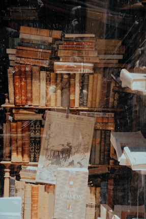 A large collection of vintage books is stacked on top of each other inside what appears to be a dusty shop window. The books vary in color, size, and thickness, with some having ornate and decorative spines. A faded, old newspaper or poster is displayed among the books, adding to the antiquarian vibe.