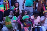 A group of people with dreadlocks are gathered together, wearing colorful clothing and accessories. Some hold flags with vibrant designs, while others have scarves and hats in red, green, and yellow. The scene has a joyful and relaxed atmosphere.