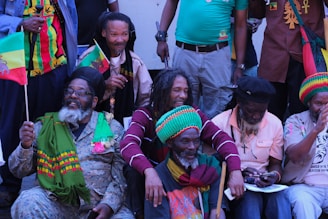 A group of people with dreadlocks are gathered together, wearing colorful clothing and accessories. Some hold flags with vibrant designs, while others have scarves and hats in red, green, and yellow. The scene has a joyful and relaxed atmosphere.