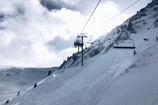 Snow-covered mountain slopes with a ski lift ascending the hillside. The sky is partly cloudy with the sun peeking through, creating a bright and serene atmosphere. Sparse trees line the mountainside, adding an element of nature to the winter landscape.