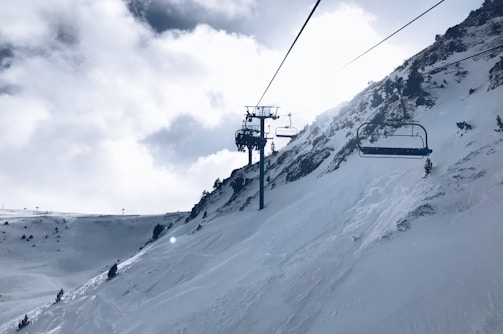 Snow-covered mountain slopes with a ski lift ascending the hillside. The sky is partly cloudy with the sun peeking through, creating a bright and serene atmosphere. Sparse trees line the mountainside, adding an element of nature to the winter landscape.