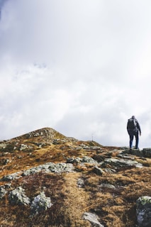 A person with a backpack is walking uphill on a rocky landscape. The terrain is composed of patches of dry grass and stones, leading to a peak with a cross in the distance under a cloudy sky.