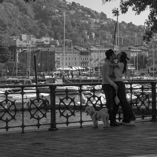 A couple embraces romantically by a waterfront promenade with a railing in the foreground. A small dog stands beside them. In the background, a marina with several boats is visible, along with hillside residential buildings. The scene has a tranquil and intimate atmosphere, enhanced by the grayscale color scheme.